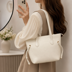 A woman in a beige and white dress carries a cream-colored textured tote bag with double shoulder straps and drawstring details, shown in a softly lit indoor setting.